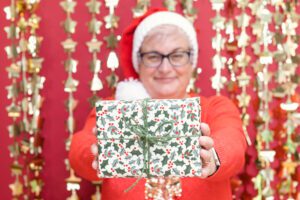 A red and gold Christmas background. Fiona is stood in a Christmas jumper and hat, holding out a wrapped Christmas present. A red and gold Christmas background. Fiona is stood in a Christmas jumper and hat, holding out a wrapped Christmas present.