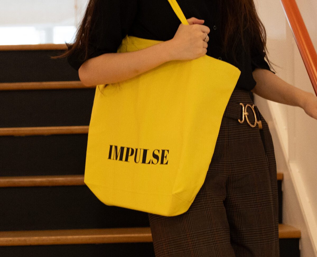 Woman walking downstairs carrying yellow tote with IMPULSE magazine logo.