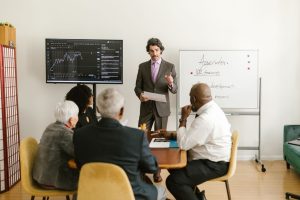 A corporate professional presents market data during a team meeting in an office setting