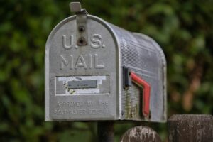 A weathered aluminum U.S. Mail mailbox with a raised red flag, mounted on a metal post beside a wooden fence, set against a blurred green foliage background.