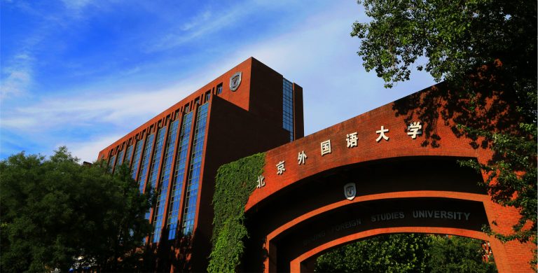 The red-brick gate and main building of Beijing Foreign Studies University under a blue sky with trees.