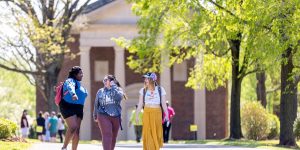 Three students walk and talk on a tree-lined path in front of a brick building at Sweet Briar College.