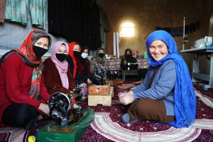 A smiling woman in a blue scarf sits with colleagues at sewing machines in a workshop, one of the Afghan women entrepreneurs supported by UNDP.