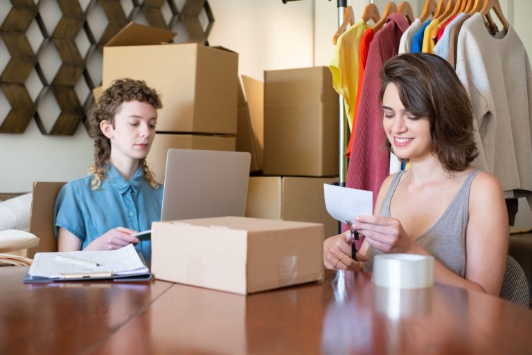 Two women manage clothing orders beside boxes and a laptop
