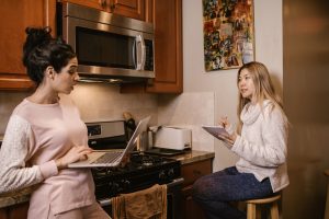 Two women collaborating in a home kitchen, using laptops and notebooks for remote work