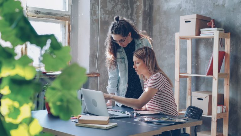 Two women collaborate at a laptop in a bright office with leafy plants